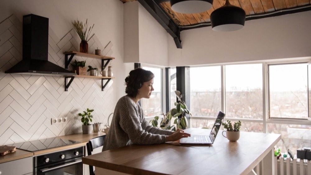 Woman working at a laptop on a wooden table in a bright apartment kitchen
