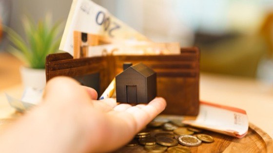 Hand holding a small model house above scattered coins