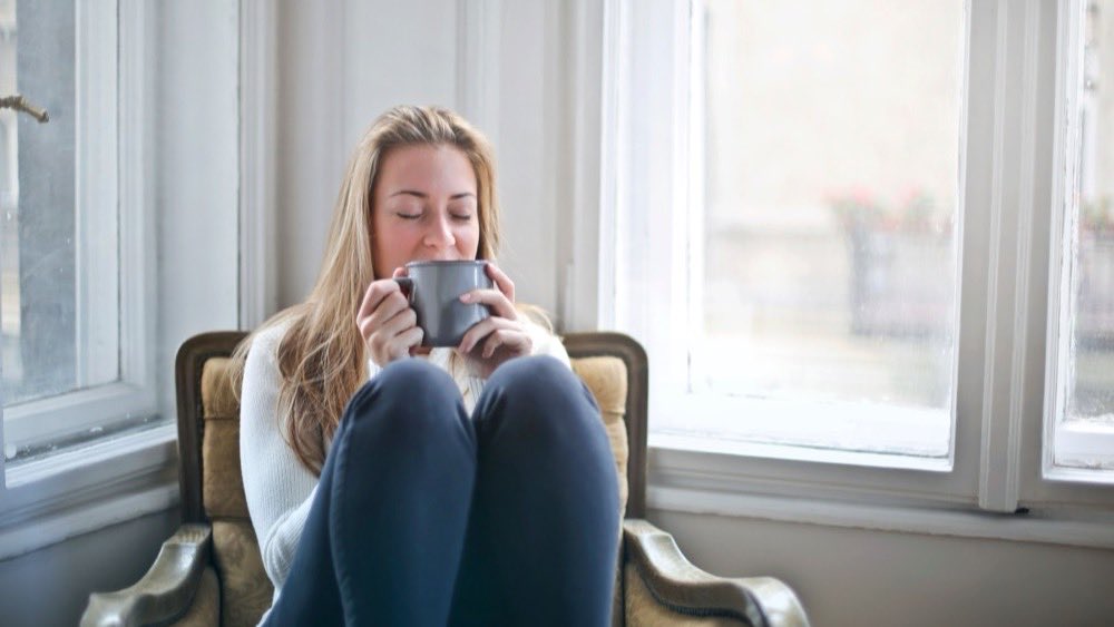 Young woman curled up in an armchair by a sunlit window holding a mug