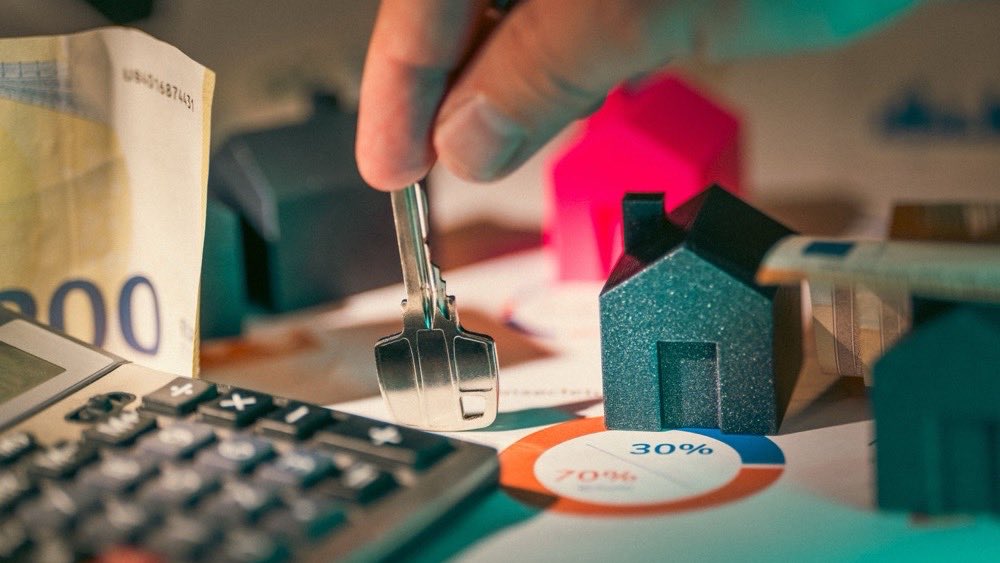 Hand placing a model house next to euro notes, coins and a calculator